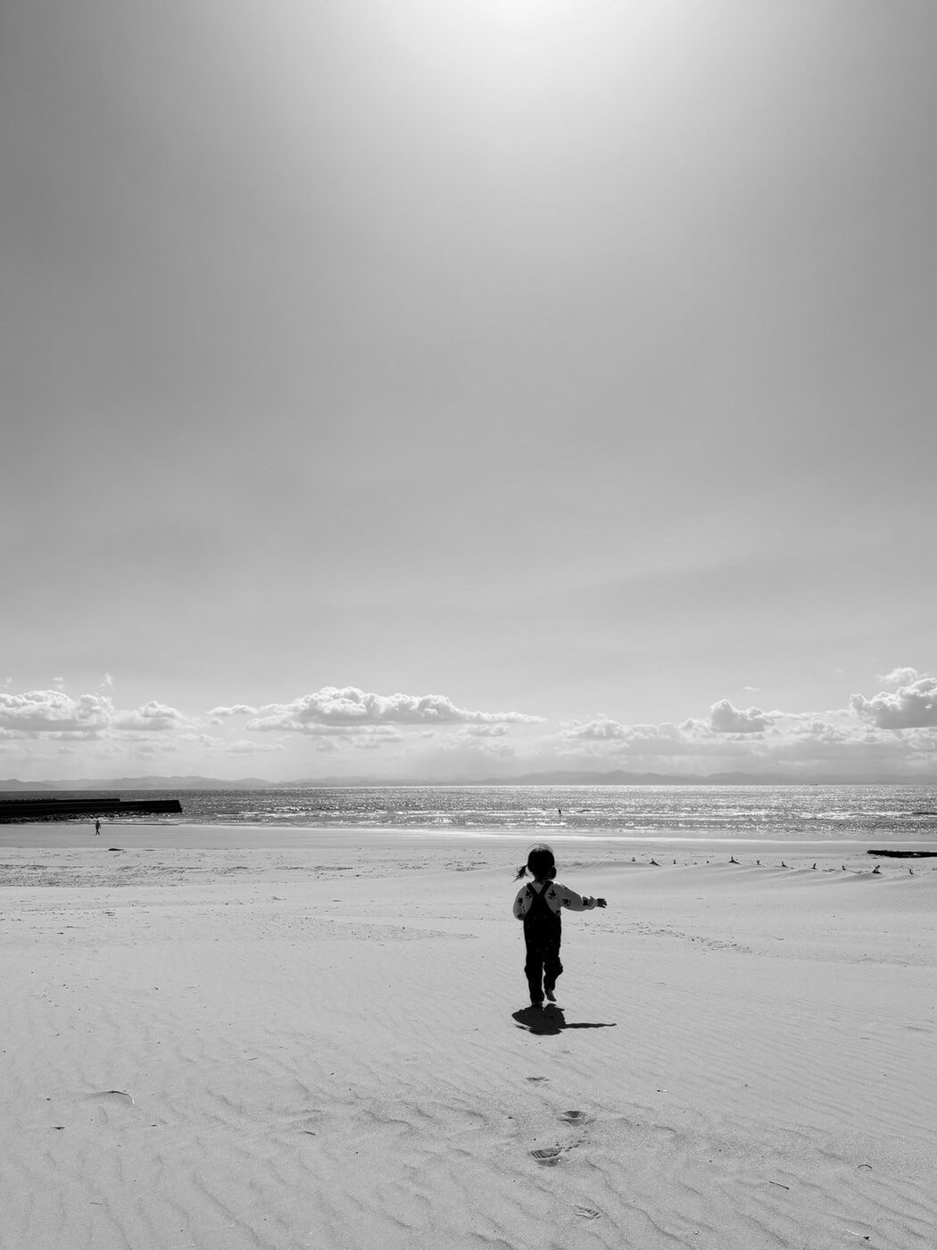 広い砂浜を歩く子どもの後ろ姿を写したモノクロ写真